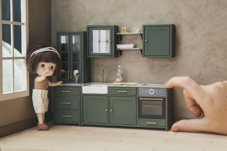 Op de afbeelding: Een miniatuur keukenset voor poppenhuizen met groene kasten, een witte gootsteen en een zwarte oven. De keuken is versierd met gouden beslag en een kleine pop staat ernaast.