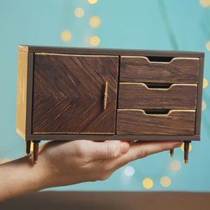 May include: A miniature wooden dresser with a single door and three drawers. The dresser is painted brown with gold accents and has small legs.