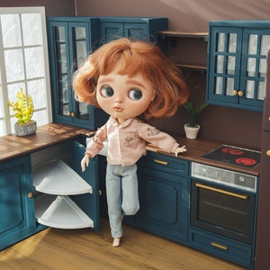 May include: A dollhouse kitchen set with a blue and white color scheme. The kitchen features a stove, oven, cabinets, and a refrigerator with the word "TED" on it. A doll with red hair is standing in front of the kitchen.