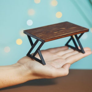 Puede incluir: Una mesa de madera en miniatura con una tapa de madera marrón oscuro y patas de metal negro. La mesa tiene forma rectangular con un diseño geométrico en las patas.