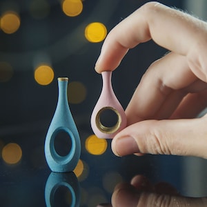 Puede incluir: Dos pequeños jarrones decorativos, uno azul claro y otro rosa claro, con detalles dorados y un recorte circular en la parte delantera. Los jarrones están sobre una superficie oscura con un fondo borroso de luces doradas.