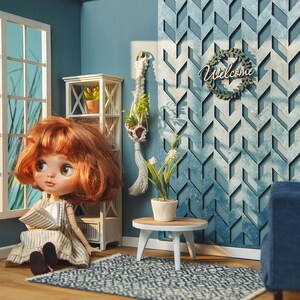 May include: A dollhouse living room with a blue and white geometric wall, a white bookshelf, a doll sitting on a rug, a small table with a potted plant, and a blue couch.