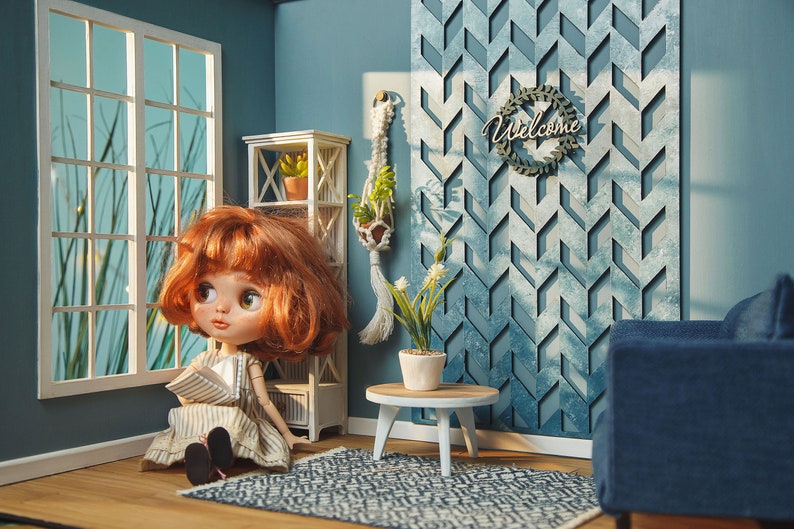 May include: A dollhouse living room with a blue and white geometric wall, a white bookshelf, a doll sitting on a rug, a small table with a potted plant, and a blue couch.