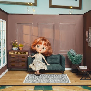 May include: A dollhouse living room with a brown couch, a doll sitting on the couch, a small dresser, and a small table with a book on it. The walls are brown and there is a window on the left side of the room.
