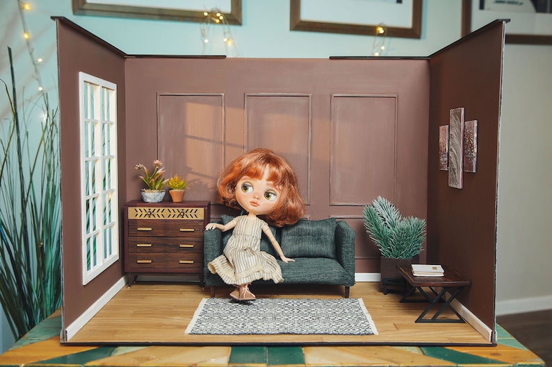 May include: A dollhouse living room with a brown couch, a doll sitting on the couch, a small dresser, and a small table with a book on it. The walls are brown and there is a window on the left side of the room.