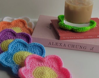 May include: Five crocheted flower-shaped coasters in various colors, including purple, orange, yellow, blue, and pink. The coasters are stacked on top of each other, with a glass of iced coffee and a book on top of the coasters.