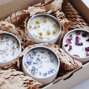 May include: Four white candle jars with lids, each filled with white wax and topped with dried flowers. The jars are arranged in a square shape in a brown cardboard box with brown paper packing material.