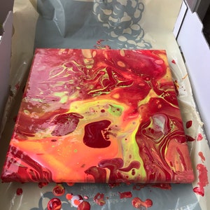 May include: Square canvas with an abstract acrylic pour painting. The artwork features a vibrant mix of red, orange, and yellow hues, with streaks of green. The paint is still wet, and the canvas is resting in a cardboard box.
