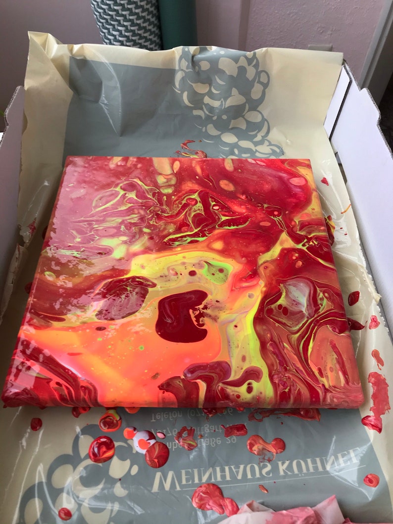 May include: Square canvas with an abstract acrylic pour painting. The artwork features a vibrant mix of red, orange, and yellow hues, with streaks of green. The paint is still wet, and the canvas is resting in a cardboard box.