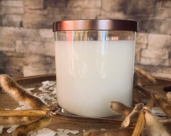 May include: A white soy wax candle in a clear glass jar with a copper lid. The candle is sitting on a wooden surface with soy flakes and dried soy pods.