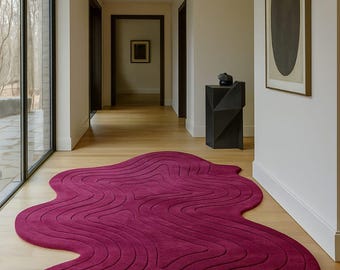 May include: A modern, abstract rug in a hallway. The rug is a deep magenta color with a flowing, organic shape and a textured, ridged pattern. The rug sits on a light wood floor.