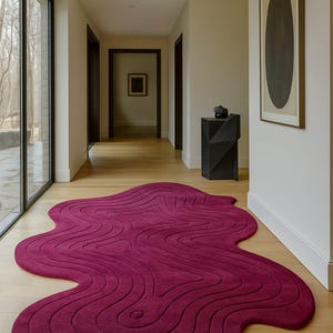 May include: A modern, abstract rug in a hallway. The rug is a deep magenta color with a flowing, organic shape and a textured, ridged pattern. The rug sits on a light wood floor.