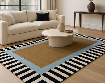 May include: A rectangular area rug with a brown center, surrounded by a light blue border and a black and white striped border. The rug is placed on a light wood floor, with a cream-colored sofa and coffee table in the background.