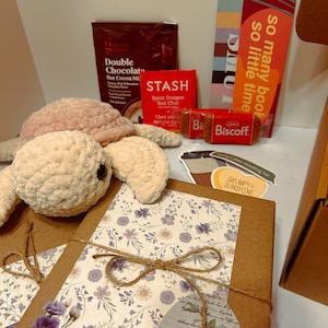May include: A gift box with a crocheted turtle, hot cocoa mix, tea, cookies, and a bookmark that says "so many books so little time." The box is decorated with floral paper and twine.
