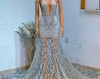 Puede incluir: Un vestido de lentejuelas plateado con un escote pronunciado y una cola larga. El vestido está hecho de una tela transparente con una superposición de purpurina plateada. El vestido está en un maniquí en una habitación blanca.