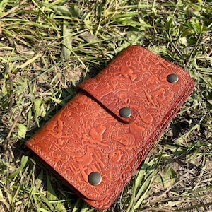 Könnte beinhalten: Eine braune Ledergeldbörse mit einem geprägten Design von Pferden und anderen Westernmotiven. Die Geldbörse hat einen Klappverschluss mit drei Metalldruckknöpfen.