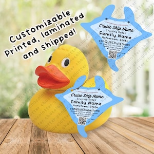 May include: A yellow rubber duck with a blue dolphin-shaped tag hanging from its neck. The tag has the text "Cruise Ship Name", "Cruising Dates", "Family Name", "Hometown, State", and "conQUACKulations".
