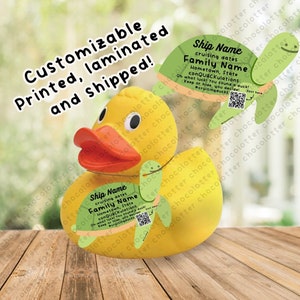 May include: A yellow rubber duck with a green turtle on a string around its neck. The turtle has a sign that says "Ship Name", "cruising dates", "Family Name", "Hometown, State", "conQUACKulations", "Oh what luck! You found a duck! Keep or hide, you decide!" and a QR code. The duck is sitting on a wooden surface.
