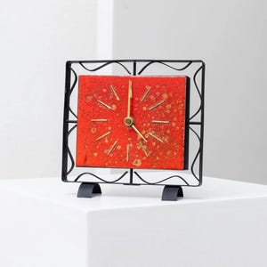 Könnte beinhalten: Eine quadratische Uhr im Mid-Century-Modern-Stil mit rotem Zifferblatt und goldenen Akzenten. Die Uhr ist in einem schwarzen Schmiedeeisenrahmen eingefasst und steht auf einem schwarzen Metallsockel. Die Zeiger der Uhr sind goldfarben.