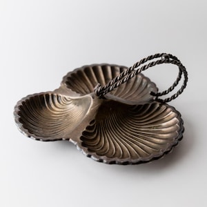 Puede incluir: Una fuente para servir de tres secciones, de color plateado, con un asa retorcida. La fuente tiene un borde festoneado y un patrón en forma de abanico en cada sección. El asa es de metal retorcido.