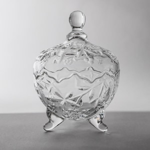 Puede incluir: Un cuenco de cristal transparente con tapa y tres patas pequeñas. El cuenco presenta un patrón detallado y texturizado y un pequeño pomo redondo en la parte superior de la tapa. El cuenco está vacío y descansa sobre una superficie gris.