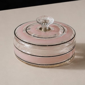 Pode incluir: Um recipiente redondo de vidro com tampa, apresentando faixas horizontais de cor rosa suave acentuadas por linhas pretas finas. A tampa é rematada com um puxador de vidro transparente e texturizado, com um design circular geral.