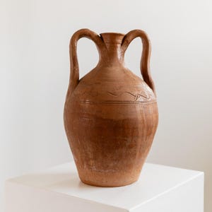 Könnte beinhalten: Eine braune Keramikvase mit zwei bogenförmigen Griffen. Die Vase hat einen breiten Boden, der sich zu einem schmaleren Hals verjüngt, mit einer dekorativen, geätzten Linie in der Mitte. Die Vase wird auf einer weißen Oberfläche ausgestellt.