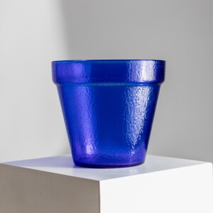 May include: A vibrant blue flower pot with a textured surface. The pot is displayed on a white cube, creating a striking contrast. The pot's shape is a classic design, perfect for showcasing plants.