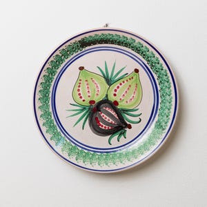 May include: A round, hand-painted ceramic plate with a fig design. The plate features a light pink rim, a green and blue border, and a central design of green and black figs with red accents and green leaves.