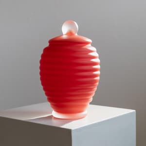 May include: A red glass jar with a ribbed design and a clear glass lid. The jar is sitting on a white pedestal.