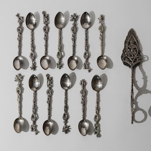 Könnte beinhalten: Eine Sammlung von zwölf silberfarbenen Metalllöffeln und einem Tortenheber. Jeder Löffel hat einen verzierten, strukturierten Griff mit komplexen skulpturalen Details. Der Tortenheber hat eine breite, spitze Klinge und einen langen, schlanken Griff.
