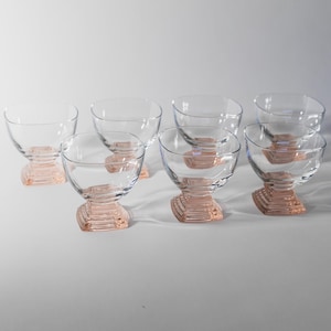 May include: A set of eight clear glass dessert bowls with a pink, geometric base. The bowls have a rounded, open top and a short, square base with a stepped design. The bowls are arranged on a white surface.