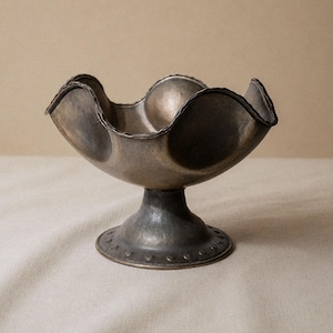 May include: A decorative metal bowl with a scalloped rim and pedestal base. The bowl has a dark, aged finish with a textured surface. The base features a series of small, evenly spaced indentations around the bottom edge. The bowl is empty and sits on a neutral-colored surface.