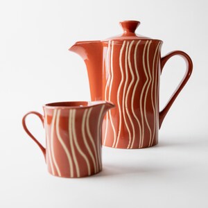 Op de afbeelding: Een terracotta-kleurige keramische koffiepot met een bijpassend melkkannetje, beide voorzien van een patroon van verticale witte golvende lijnen. De koffiepot heeft een deksel, een handvat en een tuit, terwijl het melkkannetje een handvat heeft.