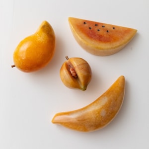 Puede incluir: Cuatro esculturas de frutas de piedra: un mango, una rodaja de sandía con semillas negras, un melocotón con hueso visible y un plátano. Las frutas son en tonos amarillos y naranjas, con una textura pulida y suave.