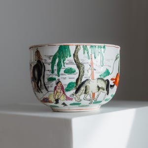 May include: A white ceramic bowl with a hand-painted scene of horses, figures, and trees. The bowl features a red rim and a detailed landscape design in green, black, orange, and other colors. The bowl is sitting on a white surface.