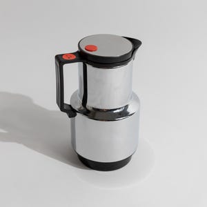 Könnte beinhalten: Eine verchromte Thermoskanne mit schwarzem Griff und Boden. Der Deckel ist silberfarben mit einem roten Knopf. Ein kleiner roter Aufkleber befindet sich am Griff. Die Kanne steht auf einer weißen Oberfläche. Für Heißgetränke.