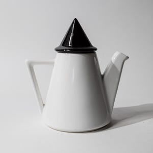Könnte beinhalten: Eine weiße Keramik-Teekanne in konischer Form mit einem glänzend schwarzen, kegelförmigen Deckel und einem geometrisch geformten Griff. Die Teekanne hat einen glatten, einfarbig weißen Körper und einen passenden weißen Ausguss in kontrastreichem Design.
