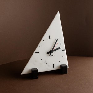 May include: A white triangular marble clock with black hands and black markings. The clock is on a brown surface and has black feet.