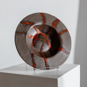 Könnte beinhalten: Eine braune Keramikschale mit kreisförmiger Form und einer zentralen Vertiefung. Die Schale weist ein auffälliges Design aus roten, abstrakten Linien auf, die von der Mitte ausgehen. Die Schale wird auf einem weißen Sockel präsentiert.