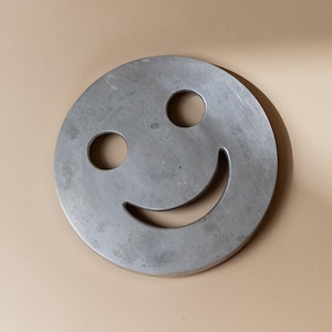 Könnte beinhalten: Großes, kreisförmiges Metallobjekt mit einem Smiley-Gesicht-Design. Das Gesicht hat zwei kreisförmige Augenausschnitte und einen gebogenen, lächelnden Mund. Das Metall hat eine gebürstete Oberfläche und ein leicht gealtertes Aussehen.