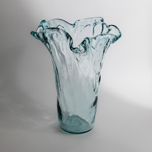 Könnte beinhalten: Eine hohe, hellblaue Glasvase mit gewelltem Rand. Die Vase hat eine strukturierte Oberfläche und eine weite Öffnung. Das Glas scheint mundgeblasen zu sein, mit leichten Farb- und Dickenunterschieden. Geeignet für Blumen oder als dekoratives Objekt.