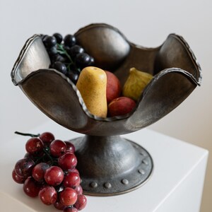 May include: A bronze-toned metal fruit bowl with a pedestal base, filled with artificial fruit. The bowl contains black grapes, a yellow pear, red peaches, and a green fig. A cluster of red grapes rests on the side.