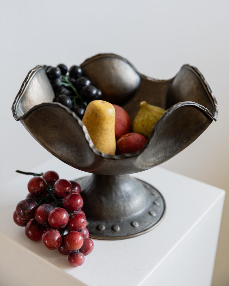 May include: A bronze-toned metal fruit bowl with a pedestal base, filled with artificial fruit. The bowl contains black grapes, a yellow pear, red peaches, and a green fig. A cluster of red grapes rests on the side.