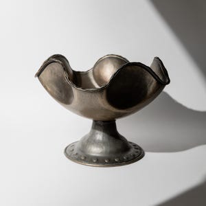 May include: A silver-toned metal bowl with a scalloped rim and a pedestal base. The bowl has a textured surface and a dark rim. The base is round with a series of small, evenly spaced rivets around the edge. The bowl is empty and sits on a white surface.