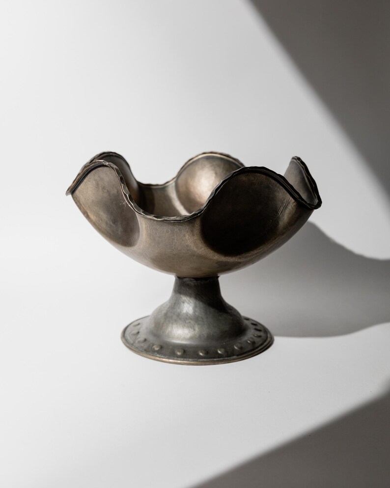 May include: A silver-toned metal bowl with a scalloped rim and a pedestal base. The bowl has a textured surface and a dark rim. The base is round with a series of small, evenly spaced rivets around the edge. The bowl is empty and sits on a white surface.