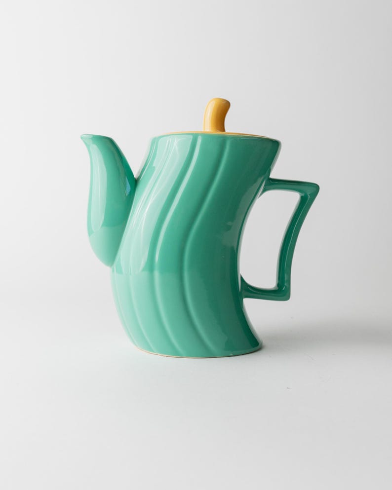 May include: A teal ceramic teapot with a curved, ribbed design. The lid is yellow, and the handle is a matching teal. The spout is curved, and the teapot has a unique, modern shape.