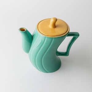 May include: A teal ceramic teapot with a yellow lid and handle. The teapot has a curved, wave-like design on the body. The handle is a dark green color.