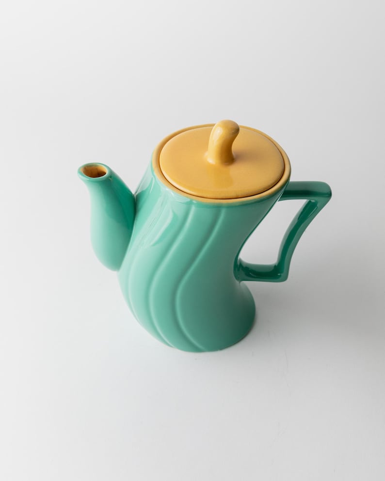 May include: A teal ceramic teapot with a yellow lid and handle. The teapot has a curved, wave-like design on the body. The handle is a dark green color.