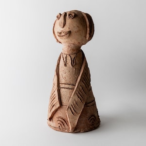 Könnte beinhalten: Eine handgefertigte, hellbraune Keramikskulptur einer stilisierten menschlichen Figur. Die Figur hat einen runden Kopf mit einfachen Gesichtszügen, einem lächelnden Mund und einem detaillierten, strukturierten Körper mit geschnitzten Linien und Mustern. Die Skulptur steht aufrecht.